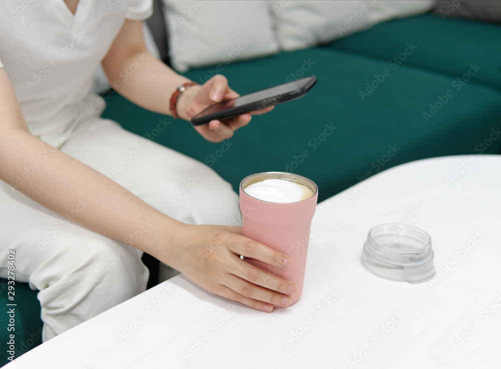 Young woman sitting on sofa looking at her mobile phone with one hand holding a pink cup full of milky drink, focused on her hand and cup. Young woman sitting on sofa looking at her mobile phone with one hand holding a pink cup full of milky drink, focused on her hand and cup.