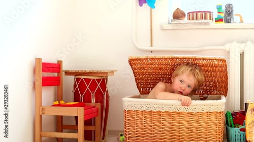 Beautiful blonde caucasian funny little male child hiding in handmade straw basket playing in kids room, smiling and looking at camera magnificent blue eyes. Happy family childhood. Modern interior.