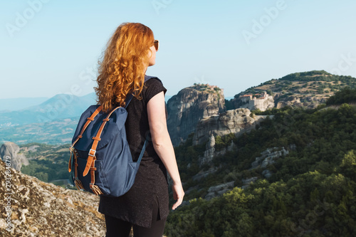 Relaxed red-haired woman tourist backpack stands on top of high mountain against the backdrop of the Meteora Mountains in Greece, and looks out over the mountains on the horizon in summer. Copy space © marina_larina