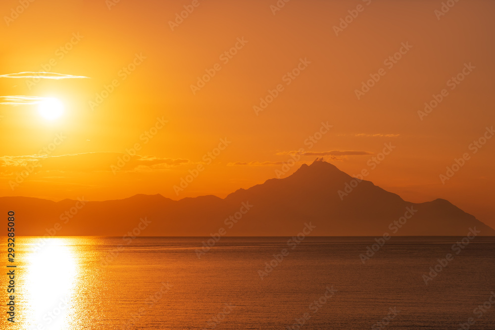 Mount Athos in Greece at sunset a bright sunrise in the early summer morning. Beautiful silhouette of Mount Athos in Greece in the Aegean Sea. Calm seawater surface. Orange disk of the sun and rays