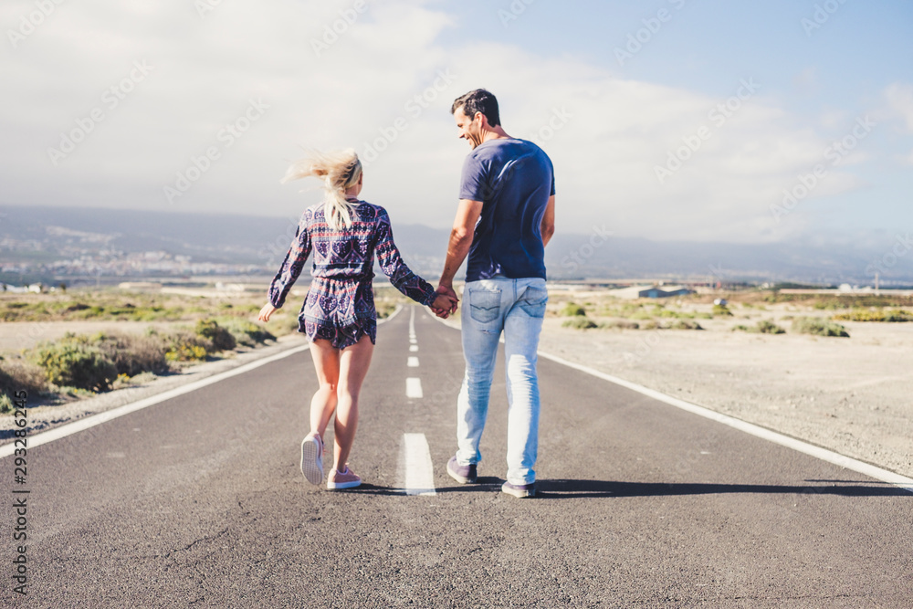 life together concept with two young people viewed from back walking together on a long straight road taking holding hands and smiling - focus on asphalt in foreground life together concept with two young people viewed from back walking together on a long straight road taking holding hands and smiling - focus on asphalt in foreground