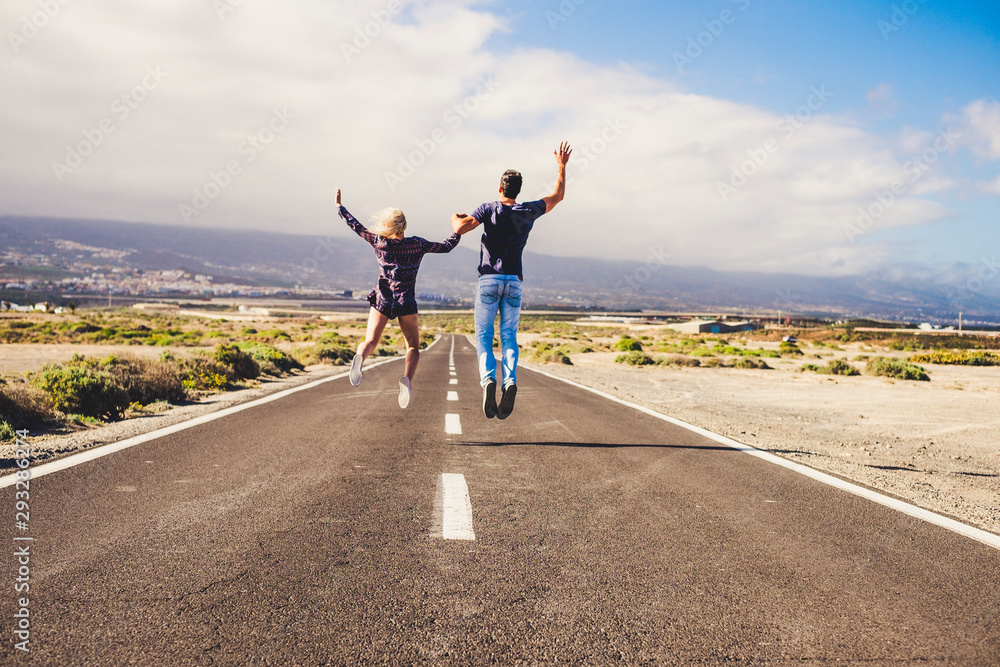 life together concept with two young people viewed from back jumping happy  together on a long straight road taking holding hands and enjoying - blue sky and mountains in background