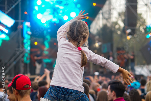 Child has fun on her parents' shoulders keeping hands with them at an outdoor rock music concert