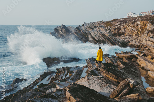 Traveler in the yellow jacket standing on the rocks in front of a huge waves