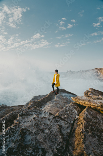 Traveler in the yellow jacket standing on the rocks in front of a huge waves