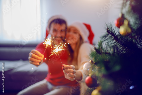 Happy caucasian couple with santa hats on heads sitting on sofa, holding sprinklers and celebrating new year. In foreground is christmas tree. Selective focus on hands.