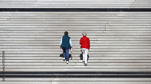 Two old women dressed in blue and red climbing up big stairs.