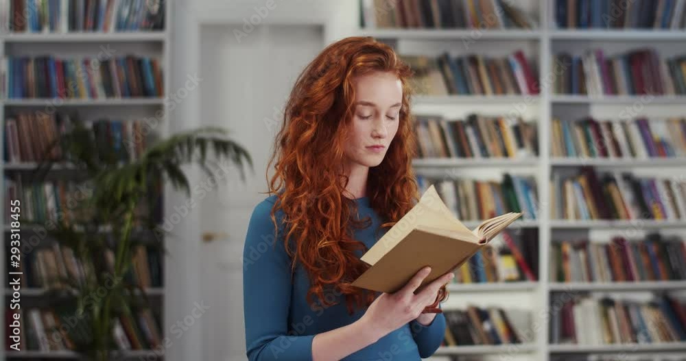 Caucasian young beautiful girl with long red hair reading an interesting story in the book while standing in the middle of the library book. Caucasian young beautiful girl with long red hair reading an interesting story in the book while standing in the middle of the library book.