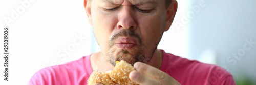 Portrait of unsatisfied male eating tasteless hamburger with obvious disgust. Bearded person holding hideous and sickening burger in hands. Blurred background