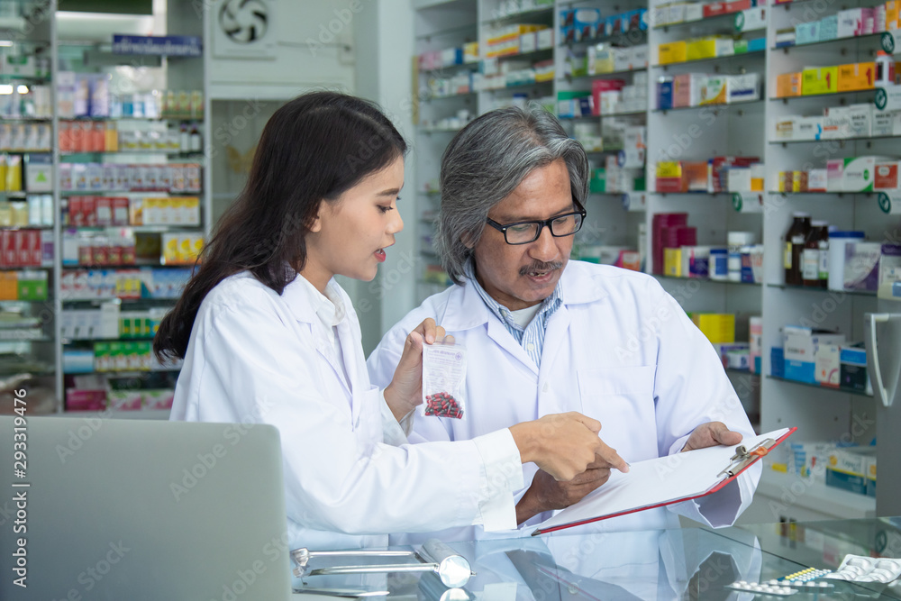 Team pharmacists colleague concentrate with pills senior man with blonde hair apothecary advice or discussing medicines with young Asian attractive colleague in drugstore blurred background