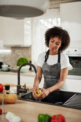 Charming mixed race woman in apron standing in kitchen ad washing yellow paprika in sink. On kitchen counter are different sorts of vegetables.