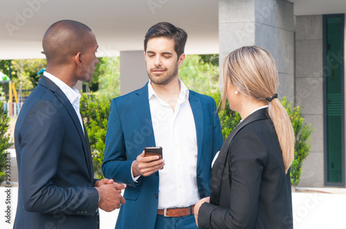 Business leader consulting mobile internet while discussing project with colleagues. Business men and woman standing near office building, using smartphone and talking. Corporate communication concept