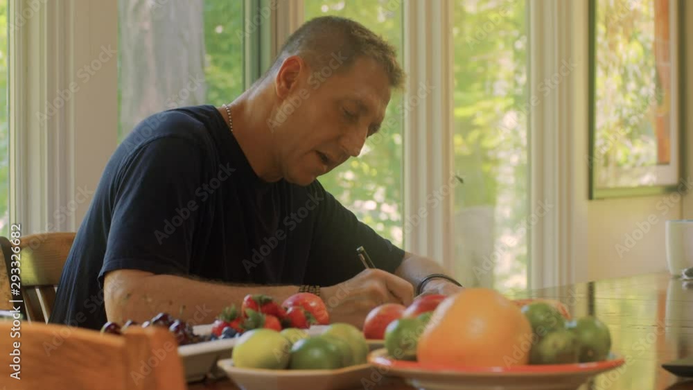 Adult man writes a note while sitting at dining room table with a fruit spread. Adult man writes a note while sitting at dining room table with a fruit spread.