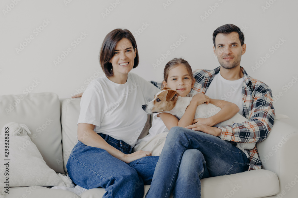 Friendly family pose together at sofa, enjoys domestic atmosphere. Father, mother, their little daughter and pedigree dog spend weekend at home, pose in living room, have happy face expressions Friendly family pose together at sofa, enjoys domestic atmosphere. Father, mother, their little daughter and pedigree dog spend weekend at home, pose in living room, have happy face expressions
