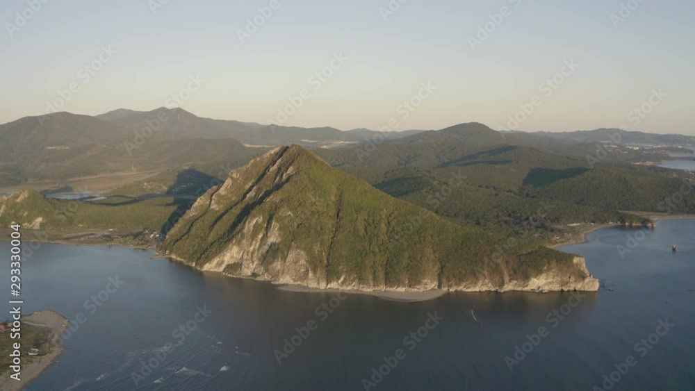 A landscape view of a pyramid-shaped mountain located at a river estuary flowing into the sea, with green vegetation on its sides, mountain ridge in the background, on the sunset. Aerial shot. A landscape view of a pyramid-shaped mountain located at a river estuary flowing into the sea, with green vegetation on its sides, mountain ridge in the background, on the sunset. Aerial shot.