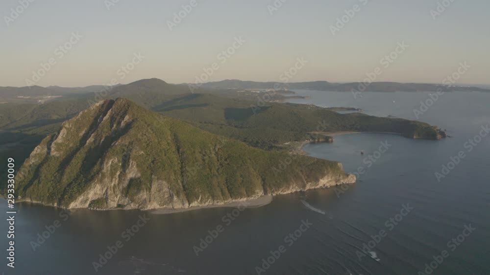 A landscape view of a pyramid-shaped mountain located at a river estuary flowing into the sea, with green vegetation on its sides, mountain ridge in the background, on the sunset. Aerial shot. A landscape view of a pyramid-shaped mountain located at a river estuary flowing into the sea, with green vegetation on its sides, mountain ridge in the background, on the sunset. Aerial shot.