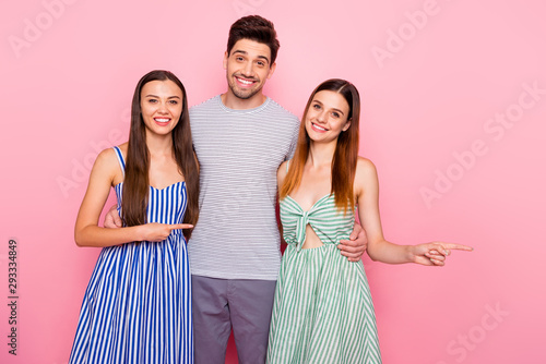 Portrait of cheerful man and two woman with long ginger foxy hair pointing at copy space wearing striped dress skirt t-shirt isolated over pink background