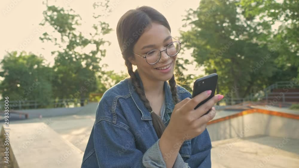 Attractive cheerful young brunette girl in denim jacket smiling and chatting on smartphone while sitting on her skateboard in skatepark Attractive cheerful young brunette girl in denim jacket smiling and chatting on smartphone while sitting on her skateboard in skatepark
