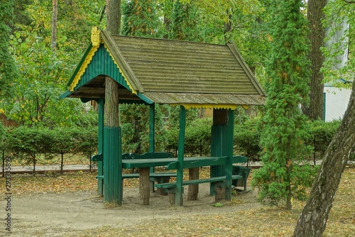 gray wooden gazebo with a green table and chairs among the vegetation and trees in the park