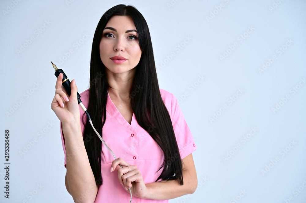 Portrait of a pretty brunette girl cosmetologist doctor, master of permanent makeup with long hair on a white background in a pink medical coat with a tool in hand. Portrait of a pretty brunette girl cosmetologist doctor, master of permanent makeup with long hair on a white background in a pink medical coat with a tool in hand.