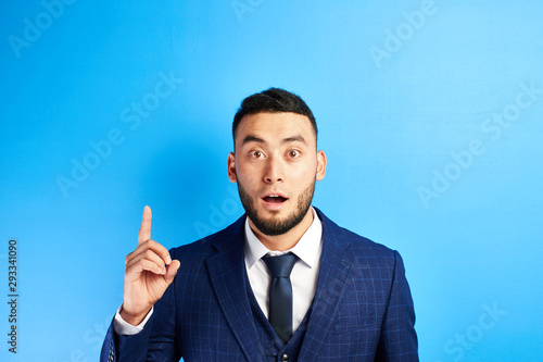 Delighted Asian Kazakh businessman has an idea, raised index finger with open mouth in a suit and tie in blue studio isolated