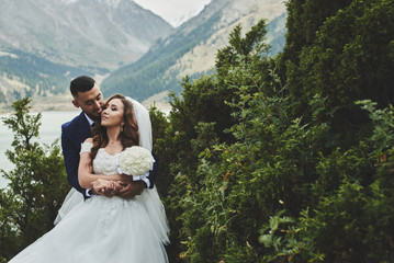 Beautiful wedding photo on mountain lake. Happy Asian couple in love, bride in white dress and groom in suit are photographed against background of the Kazakh landscape