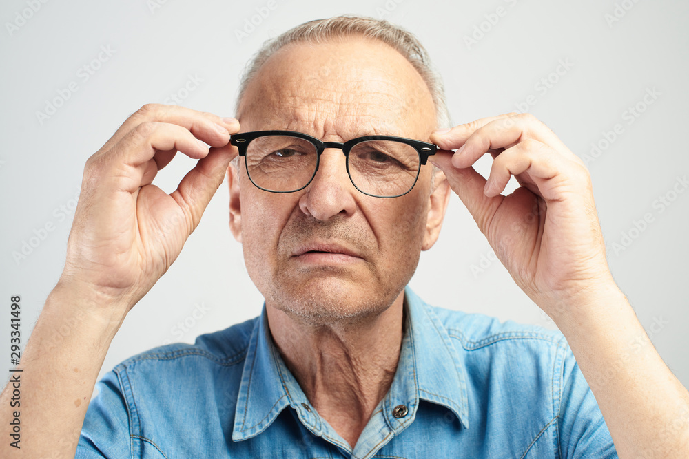 Old man in eye glasses with diopters squints, stares intently at the camera isolated on a white background. The concept of poor vision, senility blindness