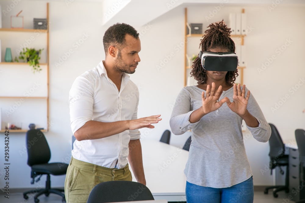 Fototapeta premium Young woman in VR headset standing in office, touching air. Her male colleague instructing her, showing hand gestures. Virtual reality simulation concept
