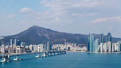 Timelapse modern bridge over azure ocean bay with motorboats turns to highway leading to Busan city skyscrapers against hilltop and clouds casting shadows zoom in photo