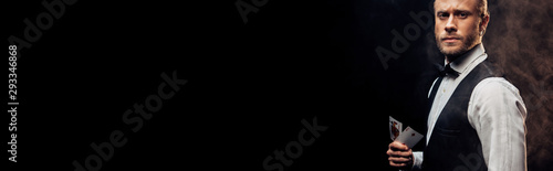 panoramic shot handsome bearded croupier holding playing cards on black with smoke