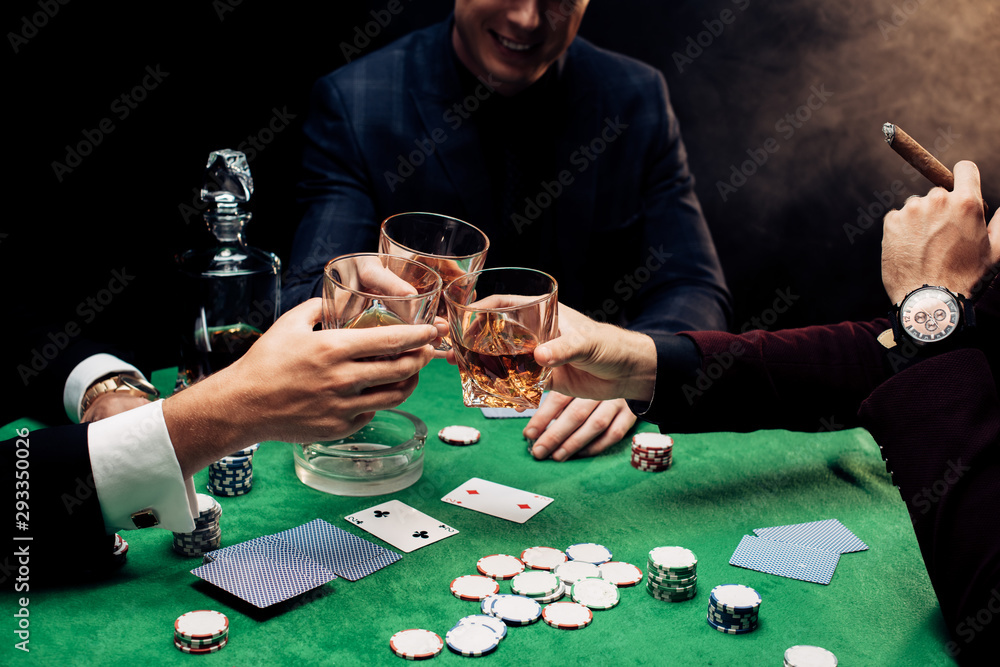 cropped view of men toasting glasses near poker table on black with smoke cropped view of men toasting glasses near poker table on black with smoke
