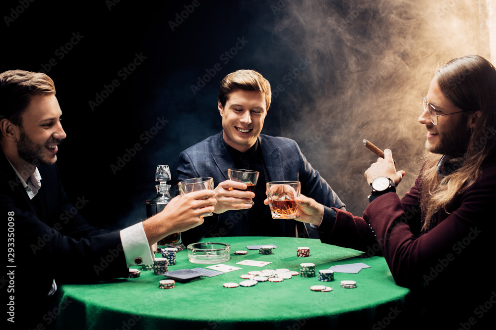 happy men holding glasses with alcohol near poker table on black with smoke happy men holding glasses with alcohol near poker table on black with smoke