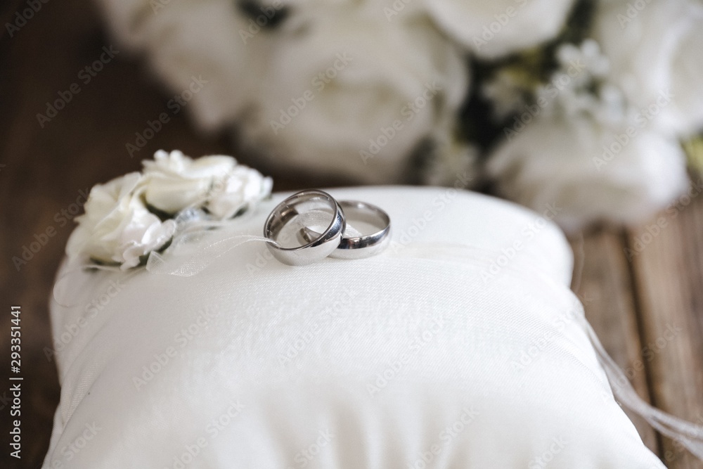 silver wedding rings on a white silk pillow with roses on a wooden surface with roses in the background silver wedding rings on a white silk pillow with roses on a wooden surface with roses in the background