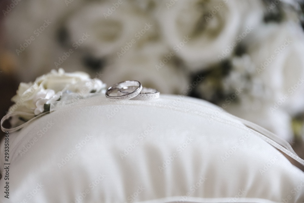 silver wedding rings on a white silk pillow with roses on a wooden surface with roses in the background silver wedding rings on a white silk pillow with roses on a wooden surface with roses in the background