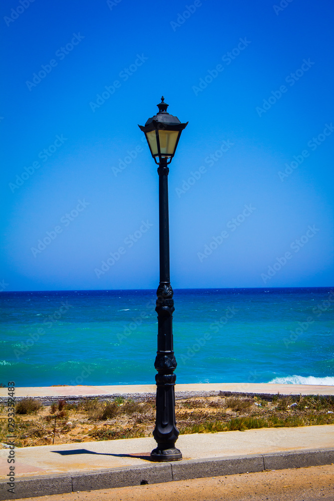 old black street lamp lantern on the beach with blue ocean and blue sky in the background