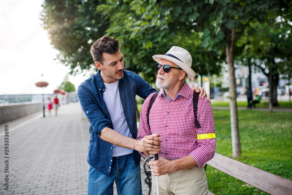 Young man and blind senior with white cane walking on pavement in city. Young man and blind senior with white cane walking on pavement in city.