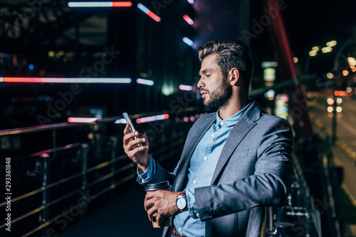 handsome businessman in formal wear using smartphone and holding paper cup in night city