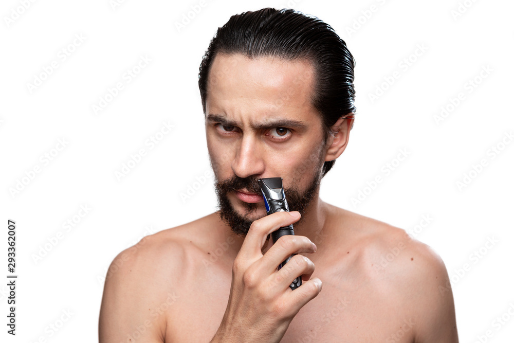 Fototapeta premium Handsome man with mustache uses electric shaver to trim his beard standning bare isolated over white background. Concept of morning treatment and shaving. Time to trim your beard. Morning routine