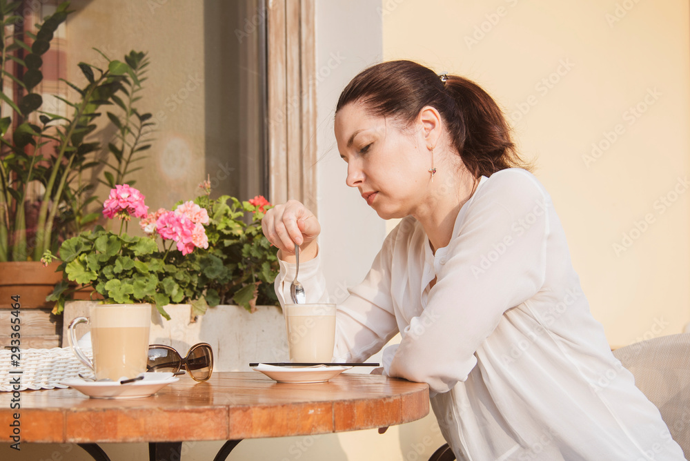 A woman sits at a table in an outdoor cafe, in front of her two cups of coffee.