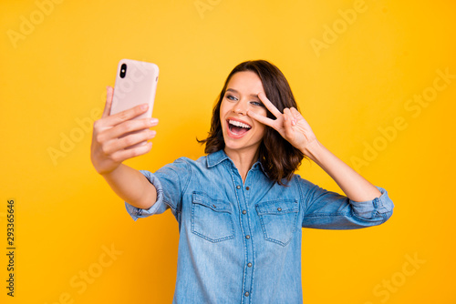 Portrait of cheerful positive girl photographer take selfie with cell phone on journey vacation make v-signs have fun feel funny funky wear denim jeans shirt isolated over yellow color background