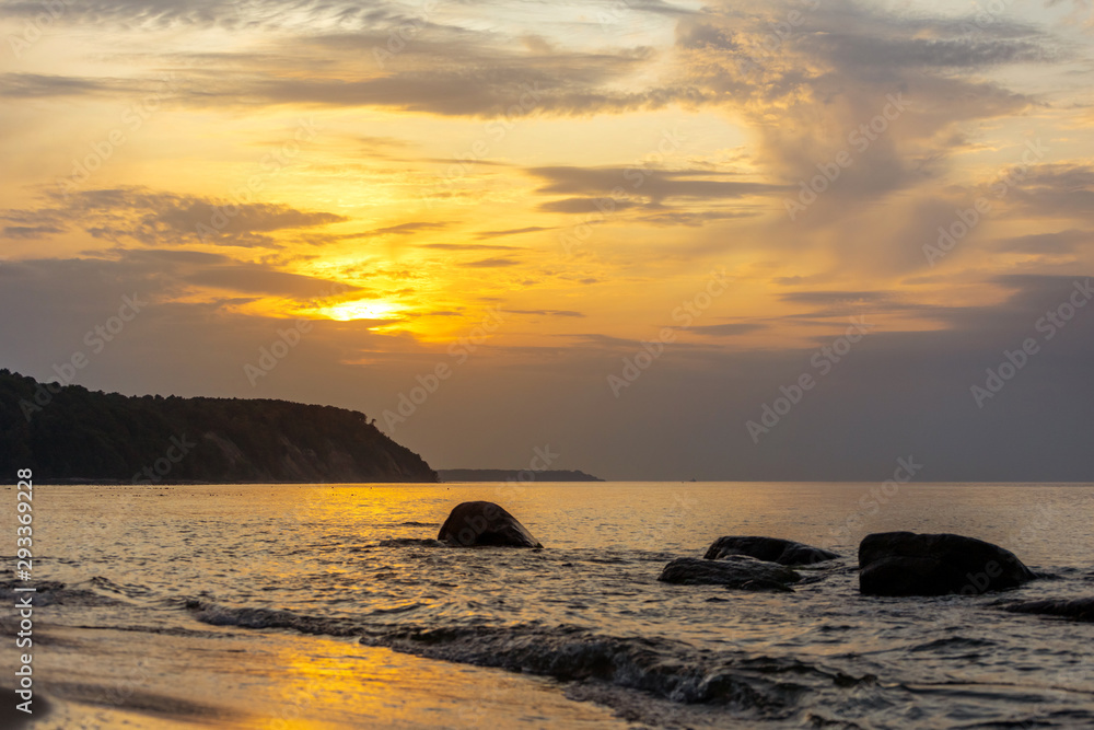 picturesque coast of the sea with a cliff in the background, large boulders in the water, low sun during sunset in orange light
