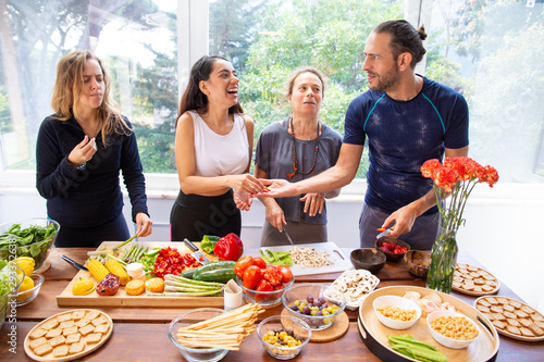 Happy friends preparing healthy meal. Cheerful young man and women in sportswear standing at table with raw organic fruits and vegetables. Healthy eating concept