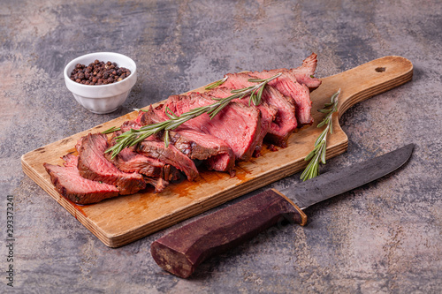 Slices of medium rare roast beef meat on wooden cutting board, old knife, pepper and rosemary twigs on gray marble background. Gourmet food. Raw meat beef steak.