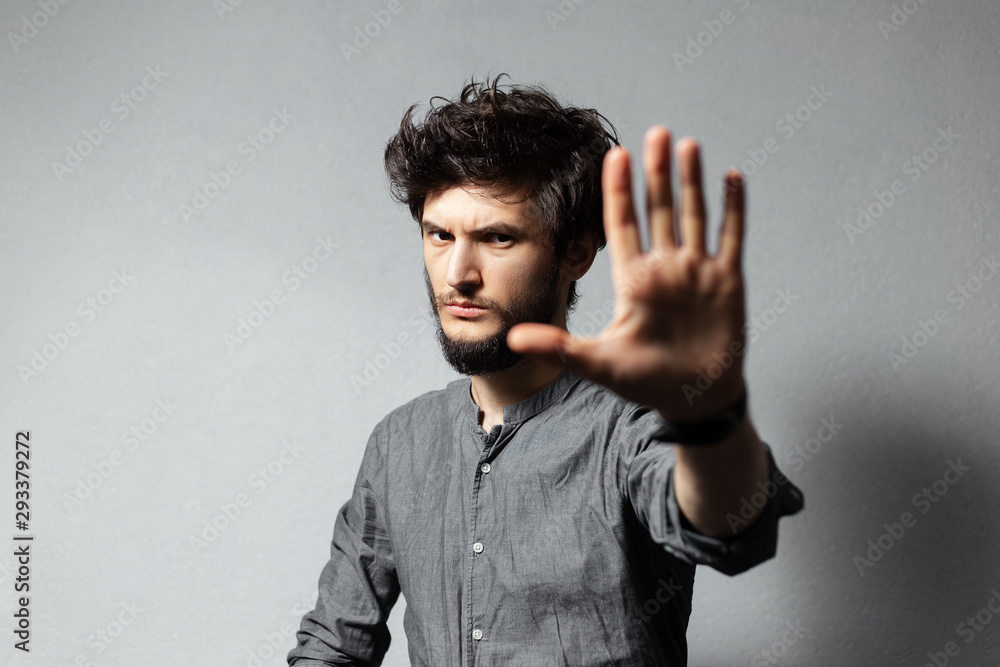 Portrait of young bearded guy with disheveled hair, shows gesture STOP with hand on background of grey. Portrait of young bearded guy with disheveled hair, shows gesture STOP with hand on background of grey.