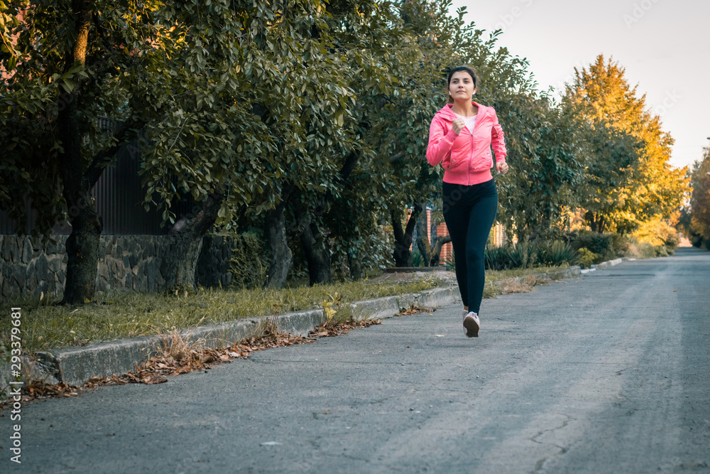 Fototapeta premium Girl runner running on sunset in autumn forest and park wearing pink jacket. Young woman jogging in fall colors. Sport and activities concept.