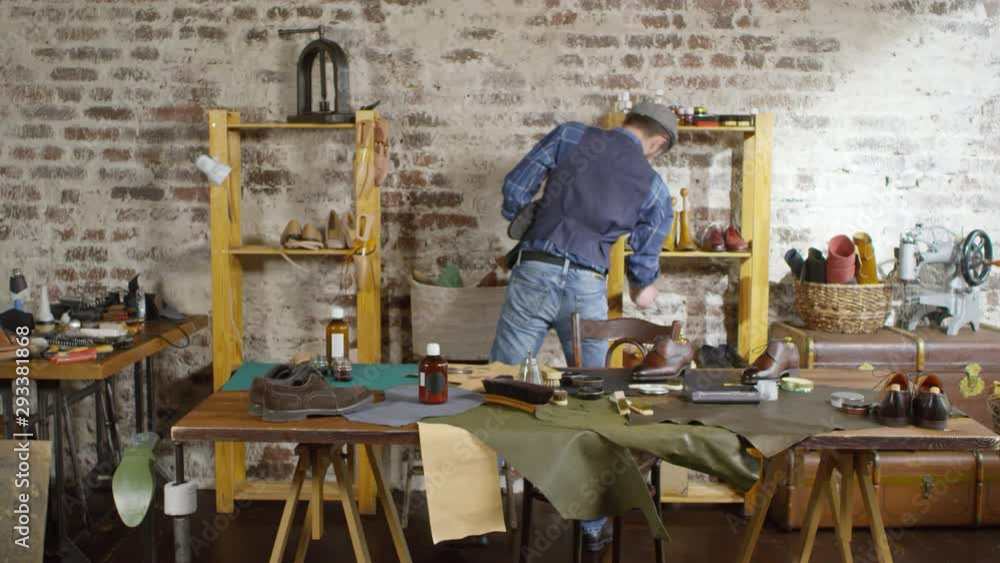 Professional male shoemaker walking into his studio, putting boots on shelves, sitting down at desk and polishing leather shoe Professional male shoemaker walking into his studio, putting boots on shelves, sitting down at desk and polishing leather shoe
