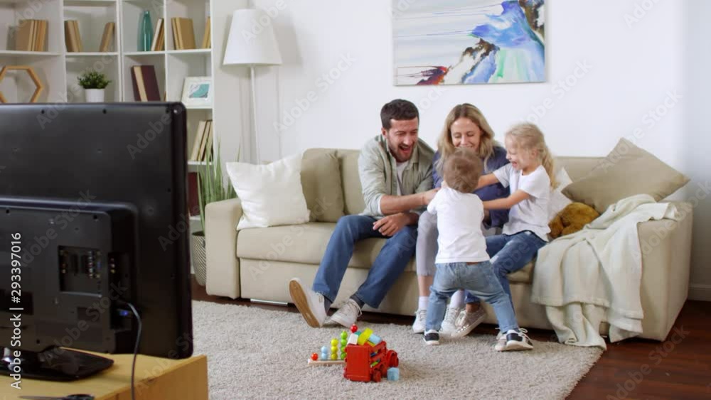 Tracking shot of bearded man and happy young woman sitting on sofa and play fighting with kids over TV remote control Tracking shot of bearded man and happy young woman sitting on sofa and play fighting with kids over TV remote control