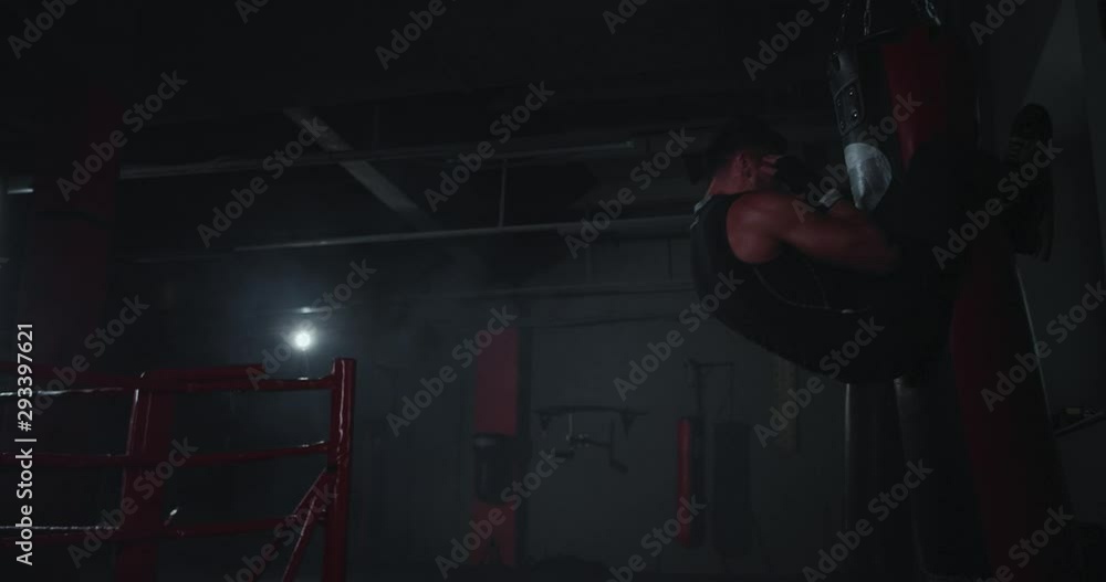 In a dark boxing class cross fit concept athletic young guy doing abs exercises while holding with legs of the boxing punching bag