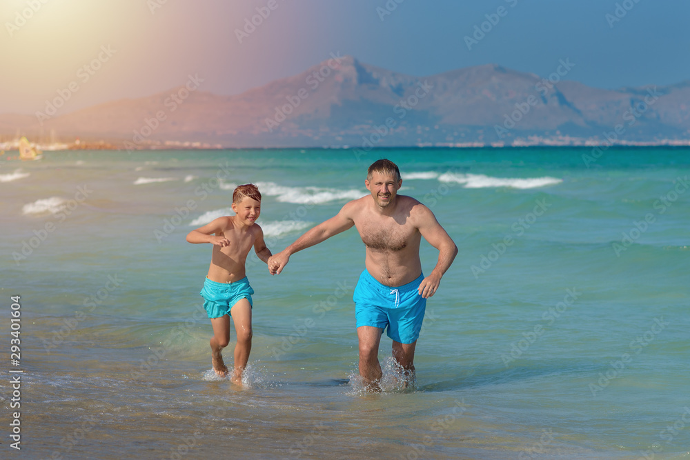 Dad and son in similar swimming shorts running along beautiful seashore. They holding hands and enjoying their perfect vacations.