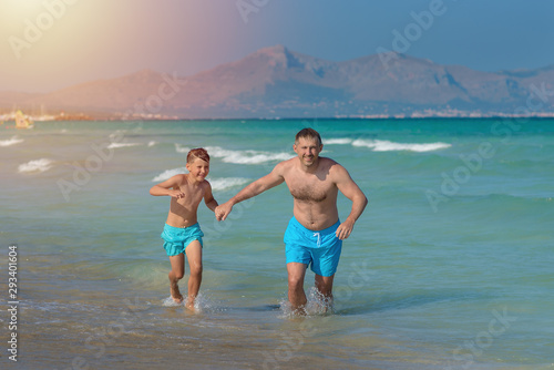 Dad and son in similar swimming shorts running along beautiful seashore. They holding hands and enjoying their perfect vacations. © Artem